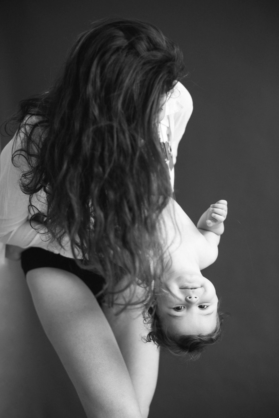 A mother playfully holds her baby upside down in a black and white studio photograph.