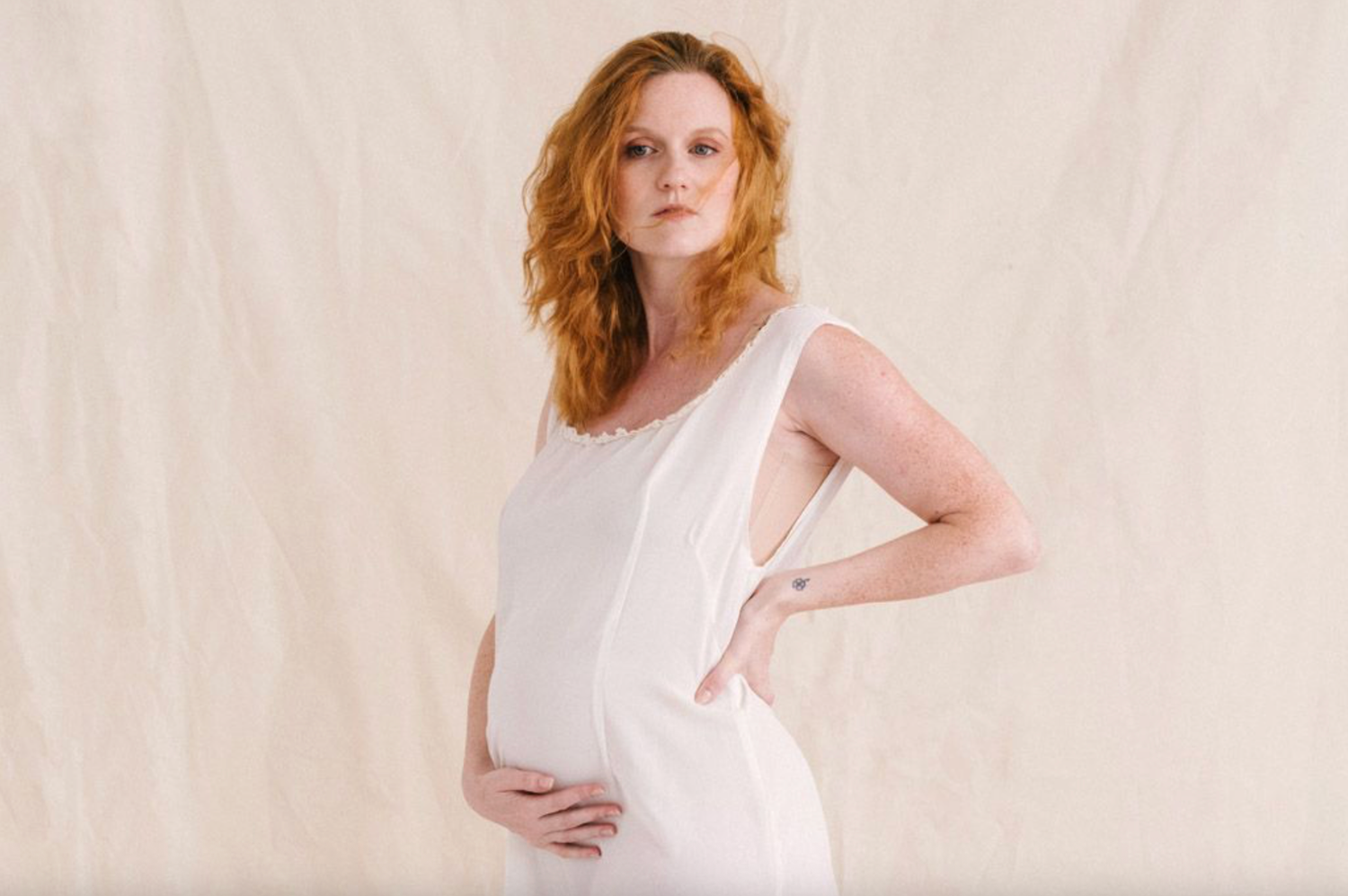 Pregnant woman in white dress holding baby bump, posing against neutral background.