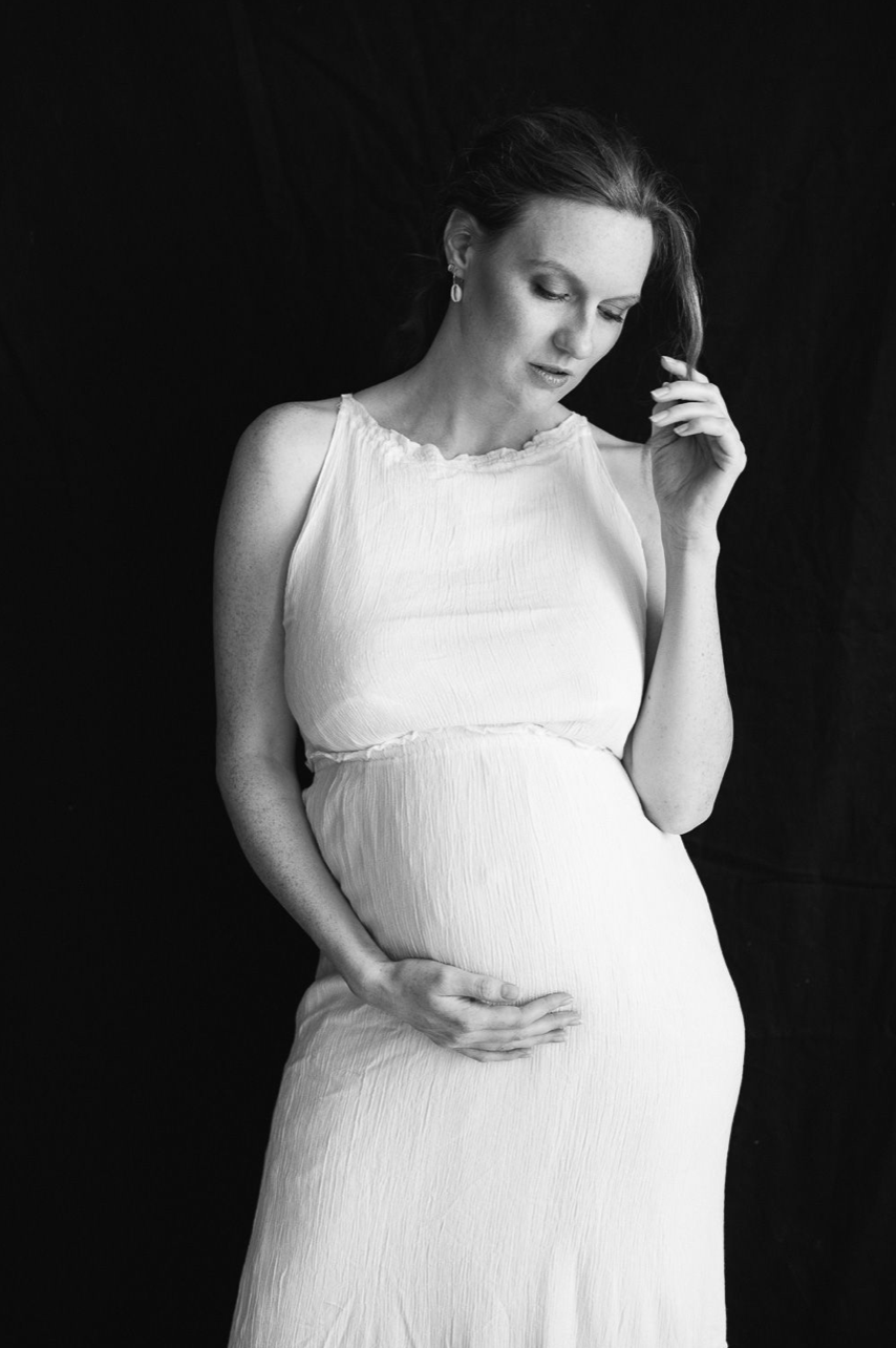 Elegant pregnant woman in white dress holding her baby bump in a black and white maternity portrait.