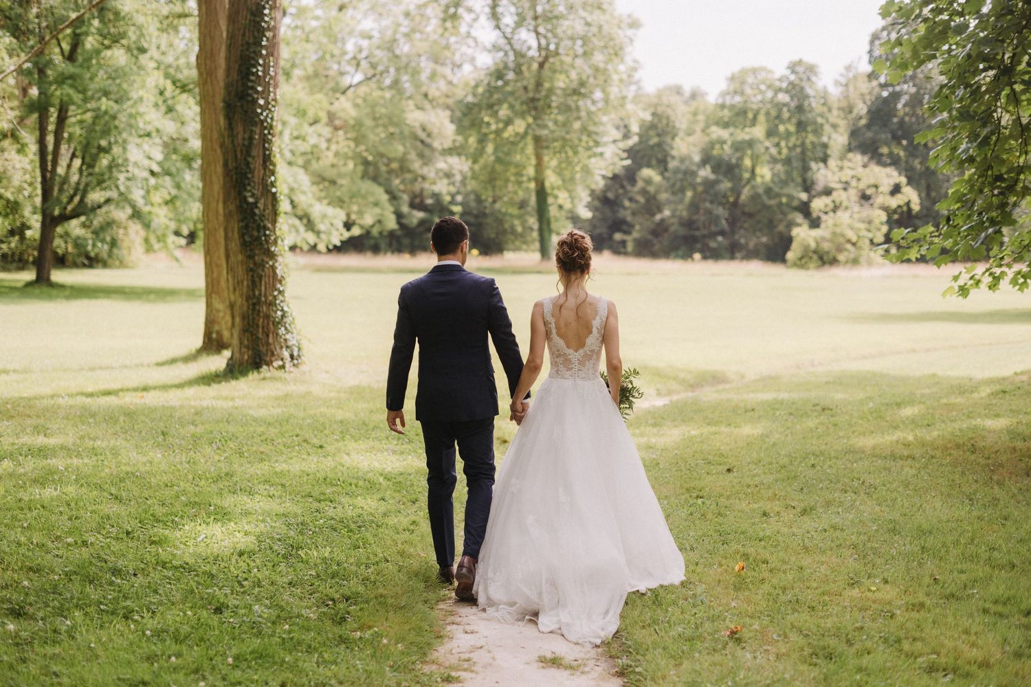Bride and groom walking hand in hand through a lush green park on their wedding day.
