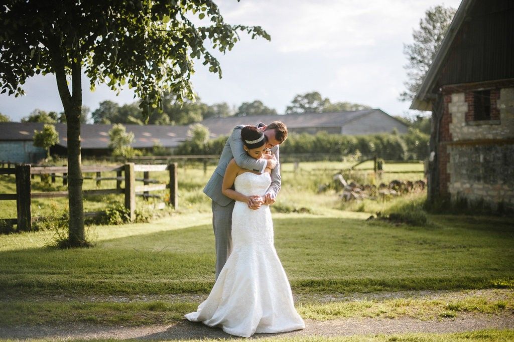 Newlyweds share romantic kiss on sunlit garden path near rustic farm buildings.