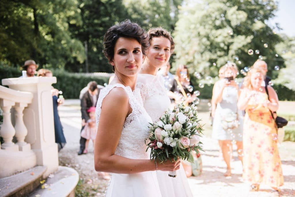 Bride in white lace gown holds pink bouquet while second bride and guests celebrate in sunny chateau garden.