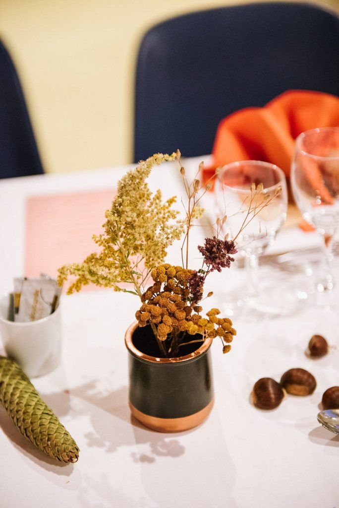 Small dark ceramic vase with copper rim holding dried wildflowers on a white wedding reception table.