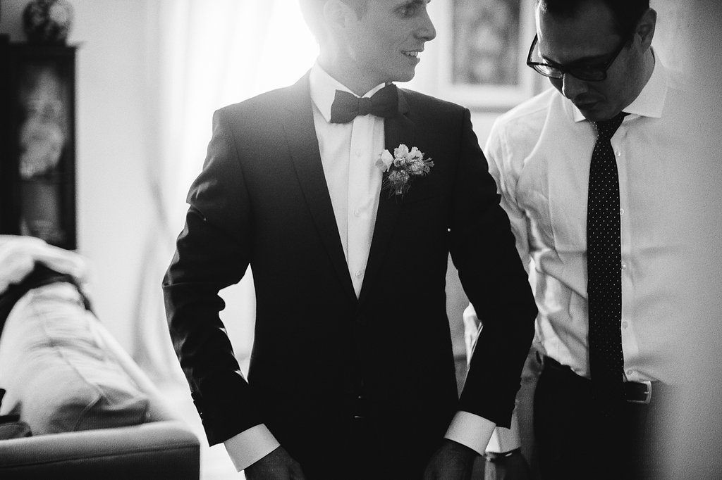 Groom in black tuxedo with boutonniere being assisted by groomsman before ceremony.