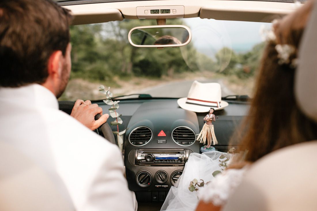 Bride and groom riding in a convertible car on their wedding day with a hat hanging from the dashboard.