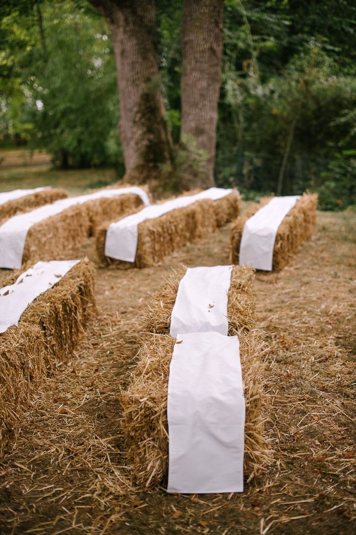 Rustic outdoor wedding seating with hay bales topped with white fabric runners arranged in rows under large trees.