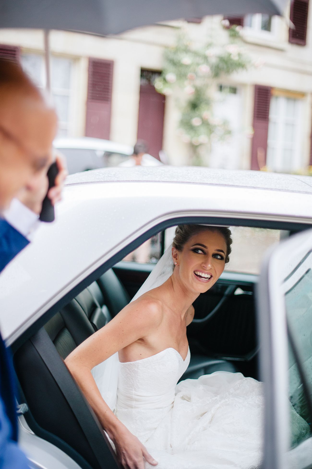 Smiling bride in strapless white wedding gown stepping out of a white car on her wedding day.