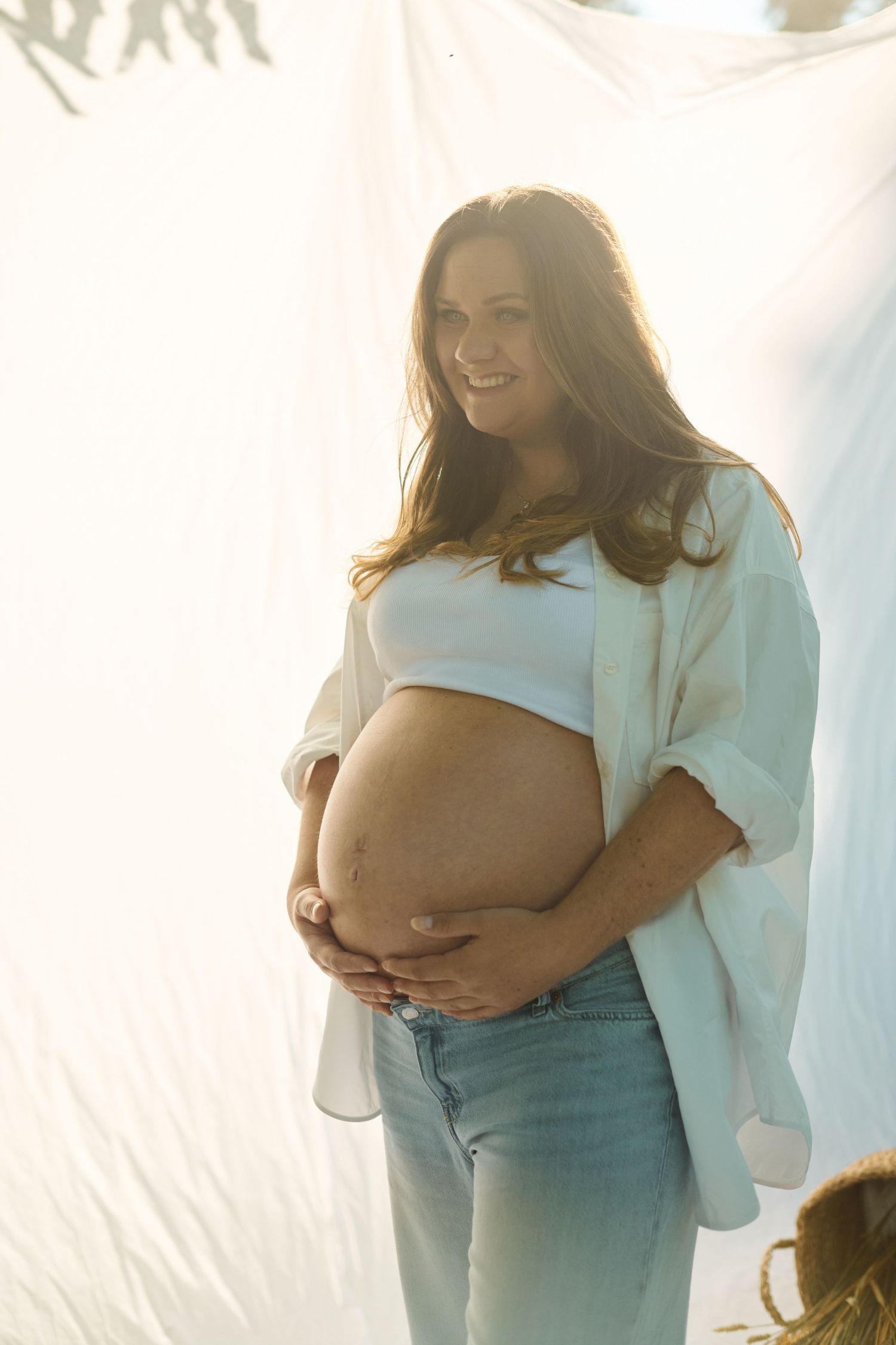 Glowing pregnant woman in white shirt and jeans holding baby bump in golden sunlight.