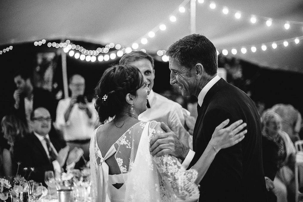 Bride and groom sharing tender moment with family member under string lights at reception.