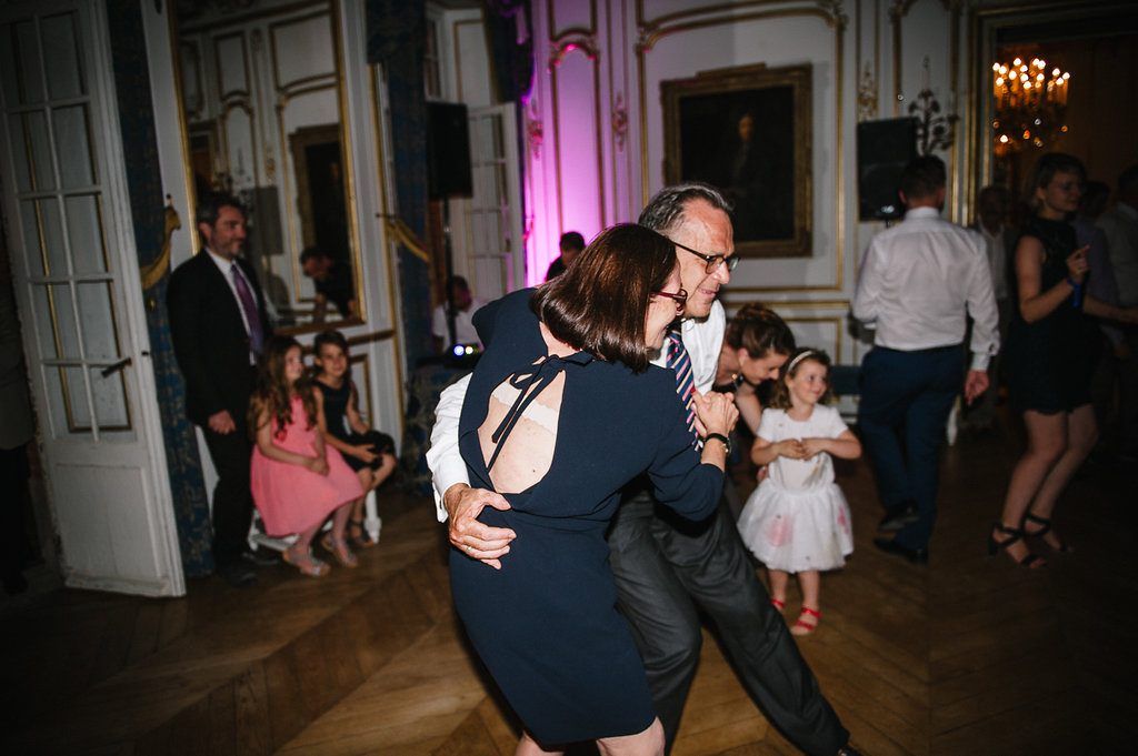 Wedding guests dance and embrace joyfully in ornate ballroom during French chateau wedding reception celebration.