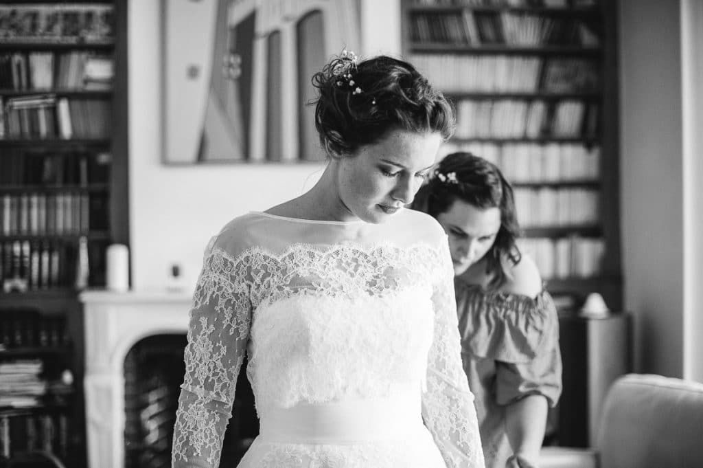 Black and white photo of a bride in lace gown being helped by a bridesmaid in a library.