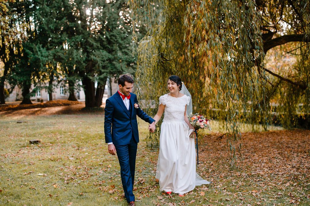 Bride and groom walk hand in hand through autumn park beneath weeping willow tree.