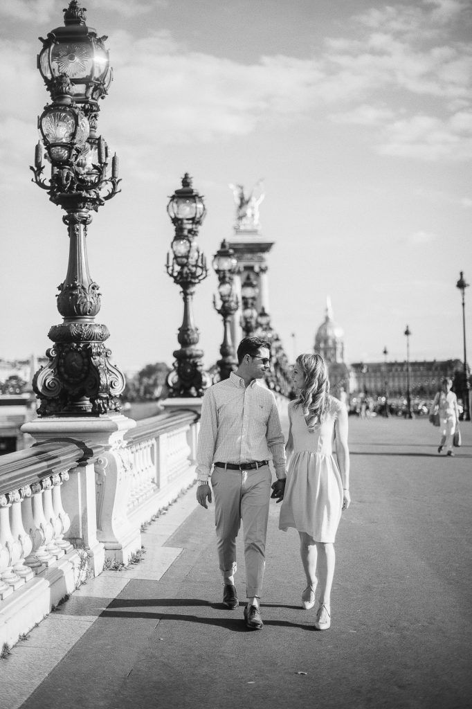 Black and white photo of a couple walking hand in hand on Pont Alexandre III in Paris.