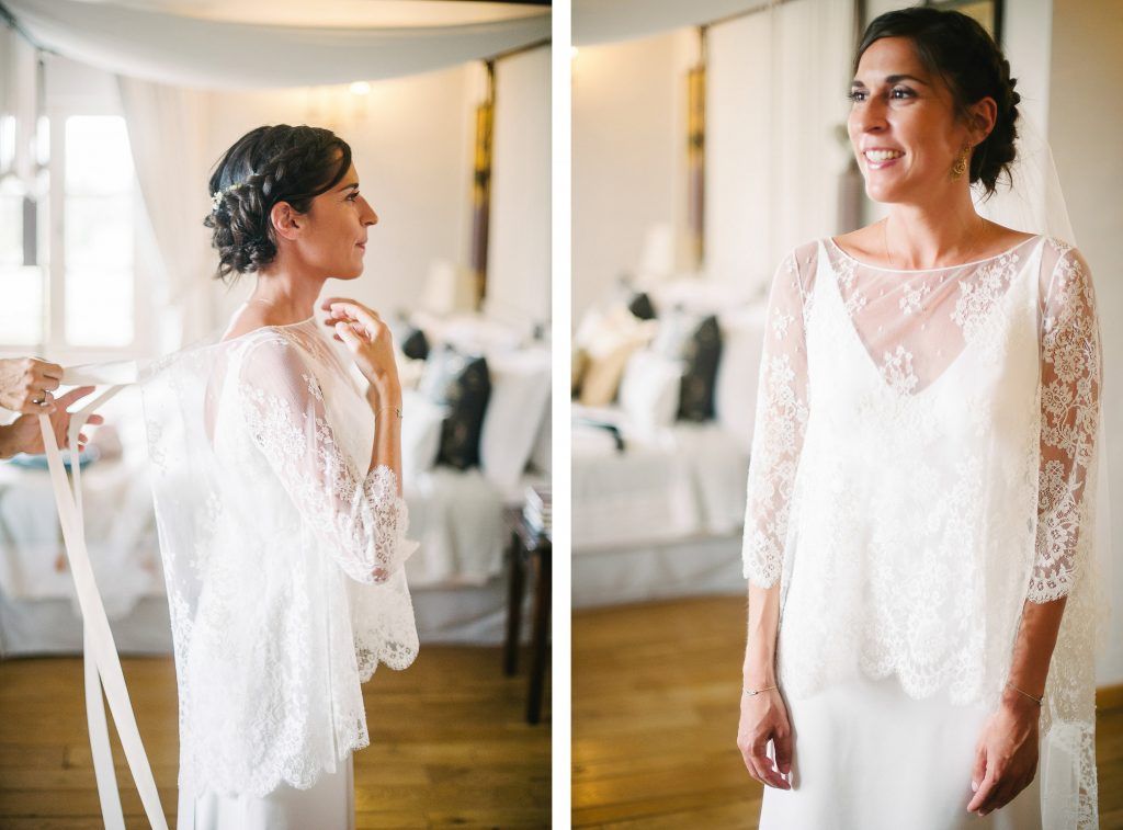 Bride wearing elegant white lace overlay wedding gown smiling in bright bedroom setting.