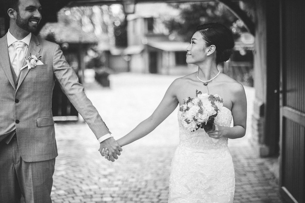 Black and white romantic photo of bride and groom holding hands, bride carrying white floral bouquet.