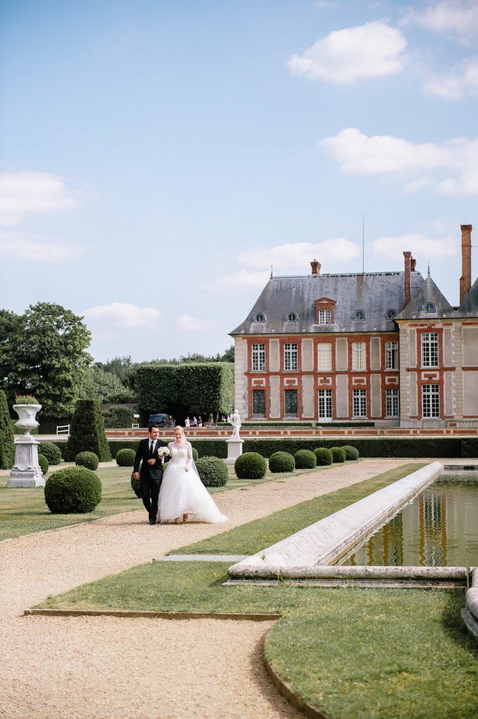 Bride and groom walk through elegant French château garden with manicured hedges and reflecting pool on sunny wedding day.