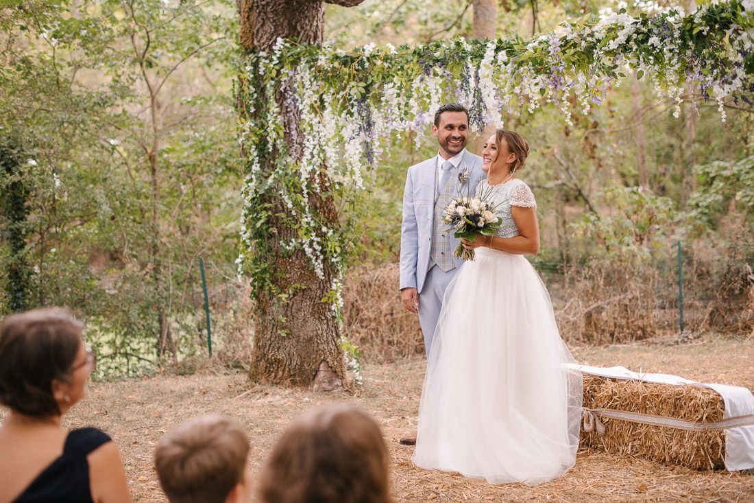 Bride and groom smiling together at outdoor woodland wedding ceremony with floral arch and hay bales.