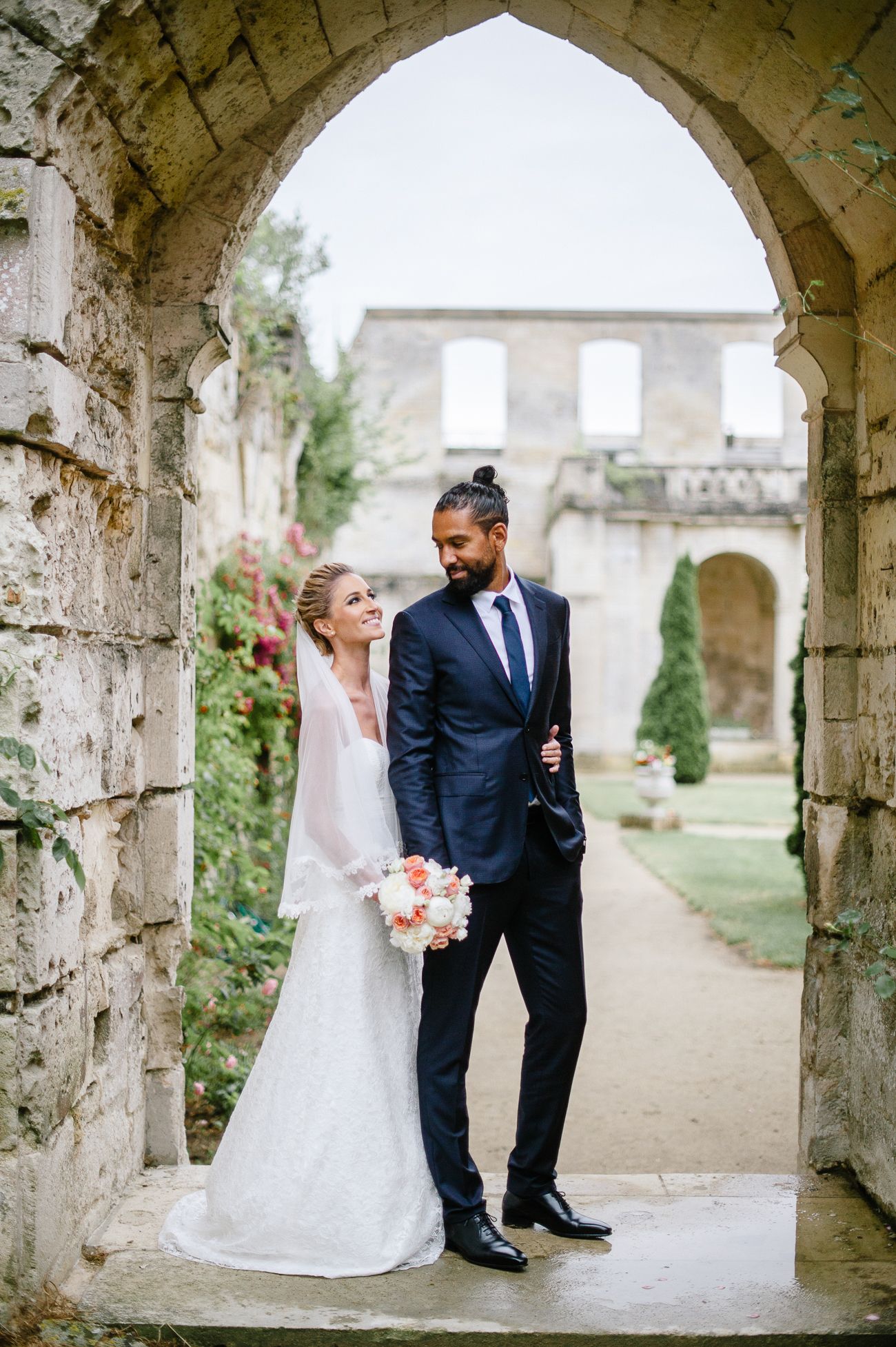 Bride and groom pose under stone arch at historic venue, bride holding pink bouquet in elegant white gown.