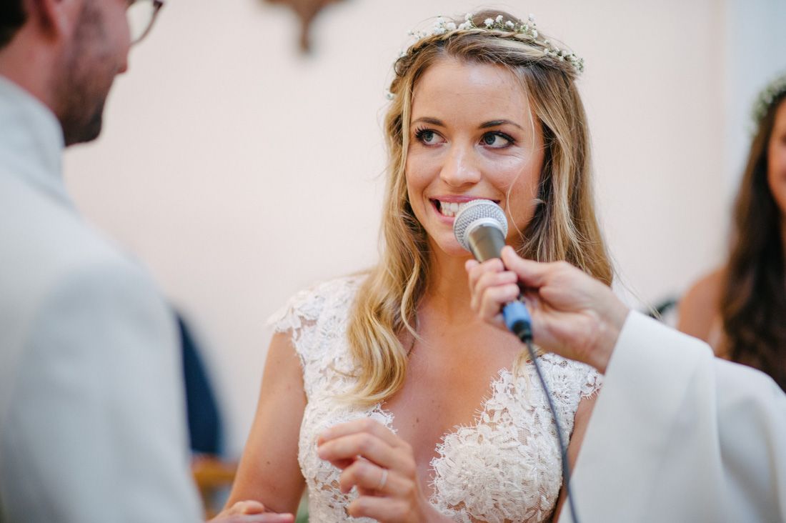 Smiling bride wearing a tiara and lace wedding dress speaking into a microphone during her wedding ceremony.