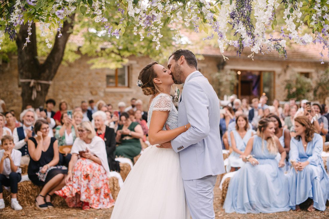 Bride and groom share first kiss under wisteria tree with joyful guests applauding.