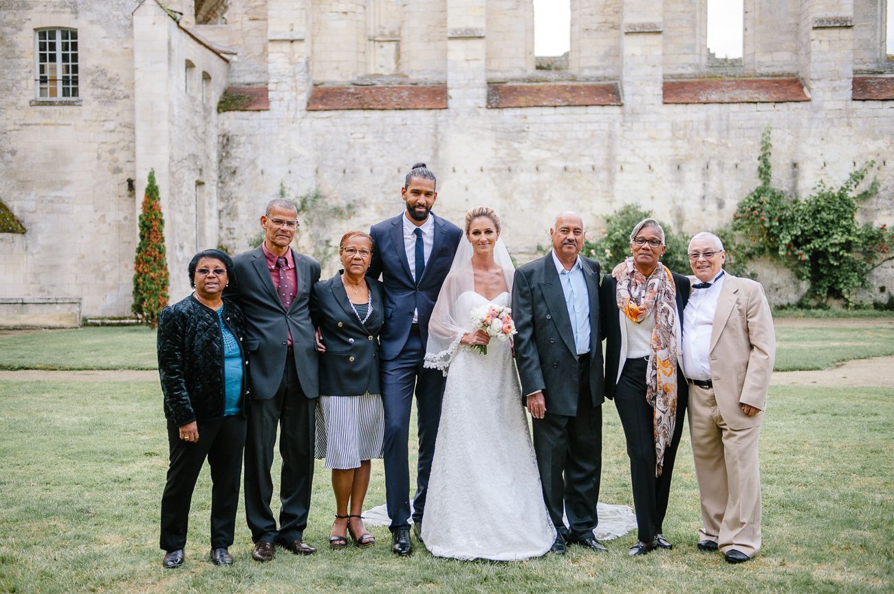 Wedding party posing outdoors on green lawn beside historic stone building, bride in white gown with groom and family.