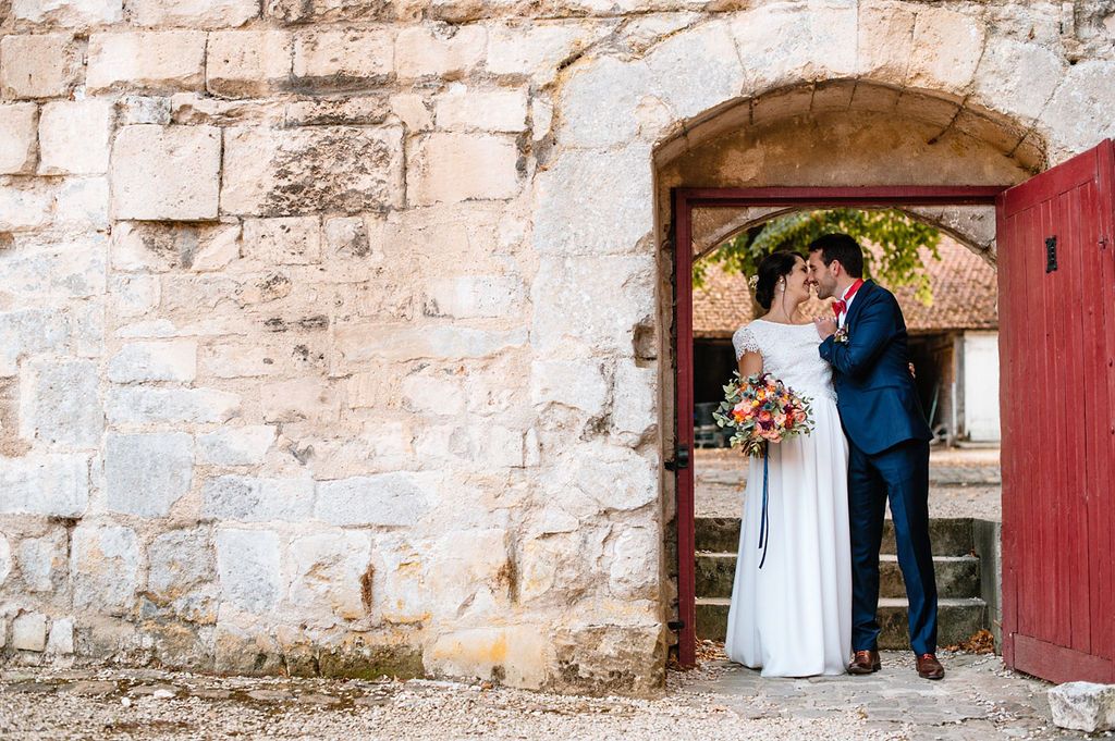 Bride and groom share a kiss framed by a rustic stone archway with a red door.