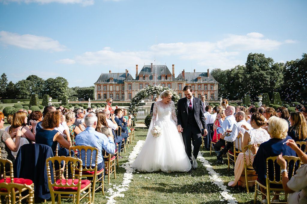 Newlyweds walk down aisle past seated guests at grand French chateau outdoor wedding ceremony.