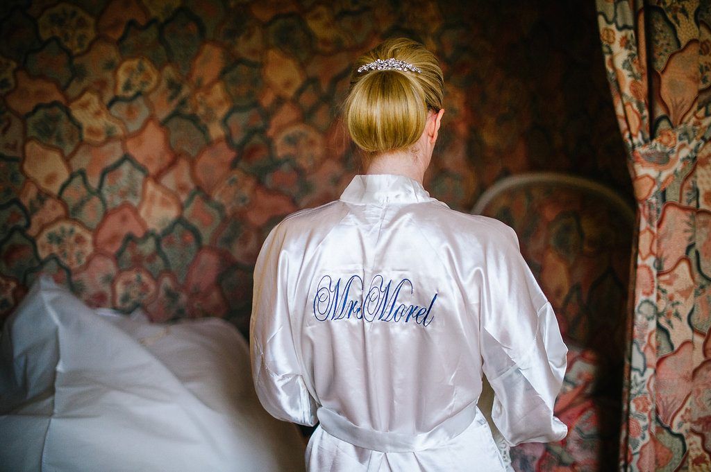 Bride wearing a white satin monogrammed Mrs. Nord robe standing in a floral wallpapered bedroom.