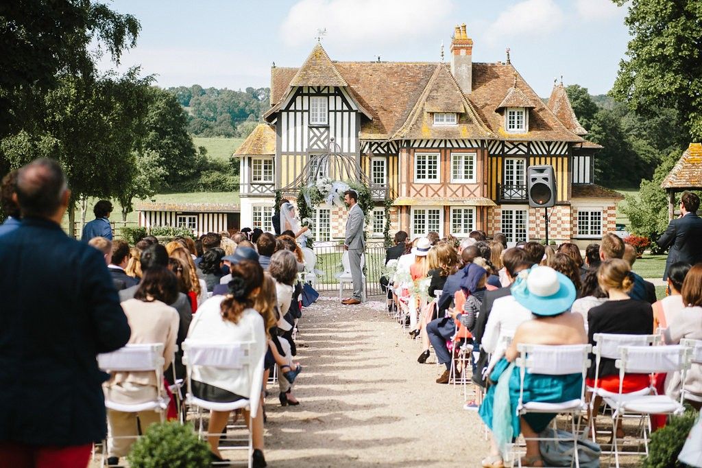 Elegant outdoor wedding ceremony at Tudor-style manor house with guests seated in white chairs.