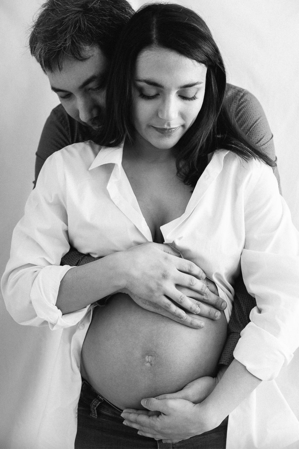 Black and white photo of a couple tenderly embracing, man hugging pregnant woman from behind holding her bump.