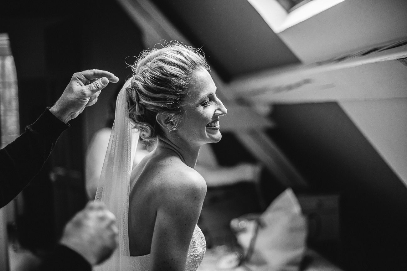Black and white photo of a smiling bride having her veil adjusted on her wedding day.