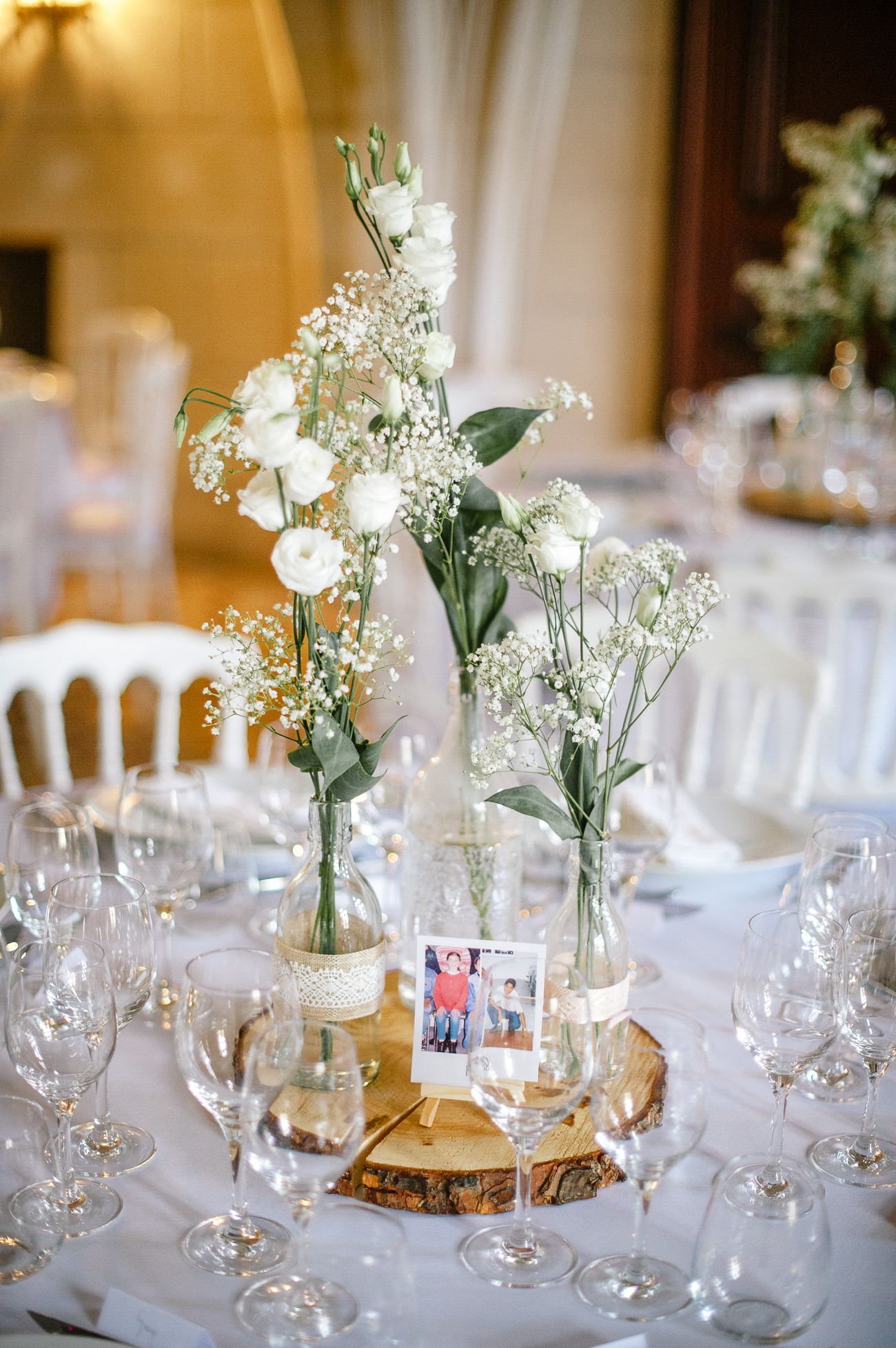 Elegant wedding table centerpiece with white roses and baby's breath in glass vases on a wood slice.