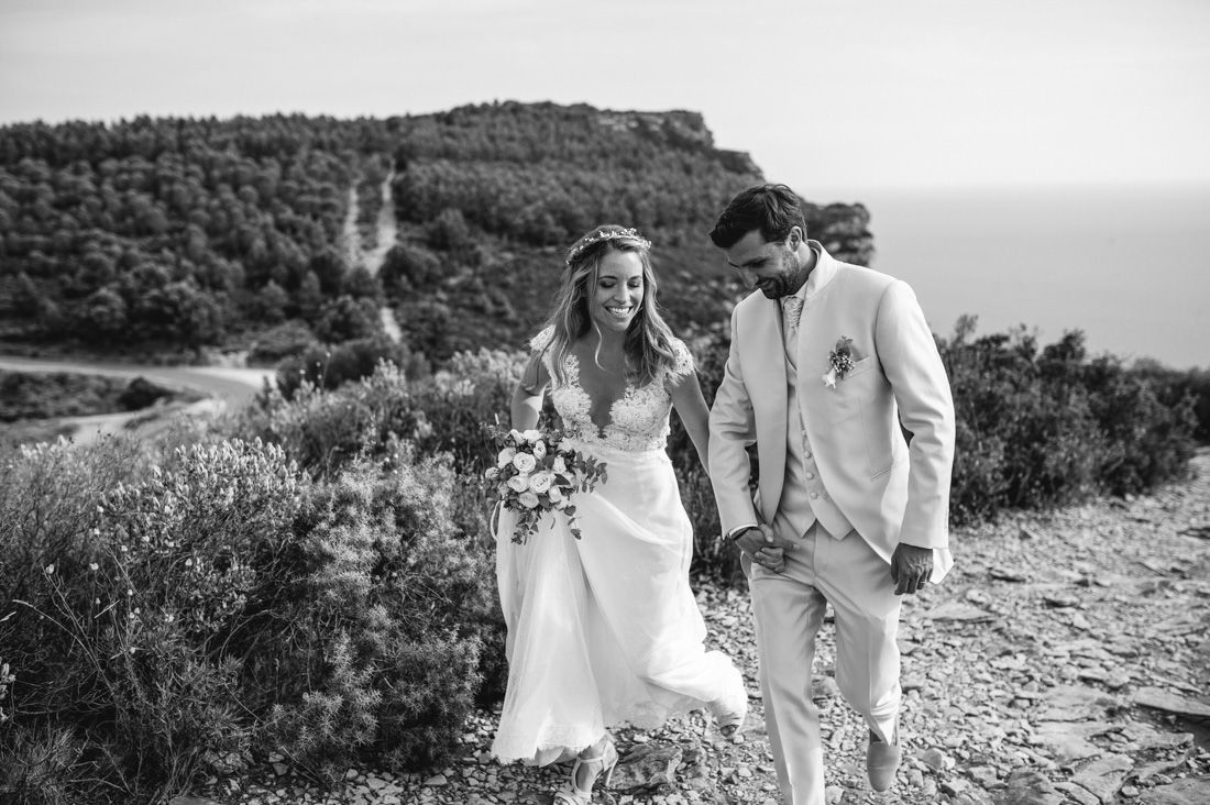 Black and white photo of a bride and groom holding hands walking along a scenic coastal hillside path.