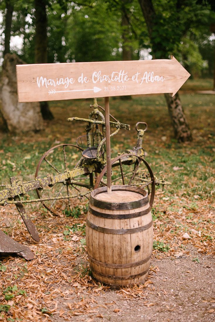 Rustic wedding sign for Charlotte et Alban on wooden barrel with vintage farm equipment in outdoor garden setting.