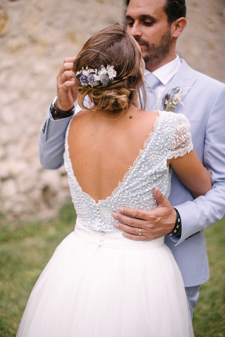 Bride in backless lace wedding dress with floral hair accessory embraced by groom in light blue suit.