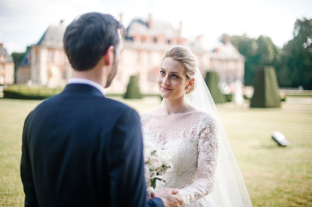 Bride in lace wedding gown gazing at groom with elegant chateau in background.