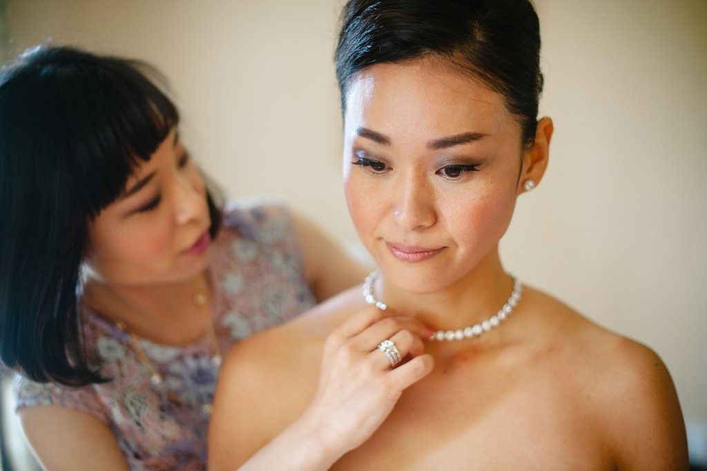 Helper clasping pearl necklace on smiling Asian bride wearing strapless wedding dress.