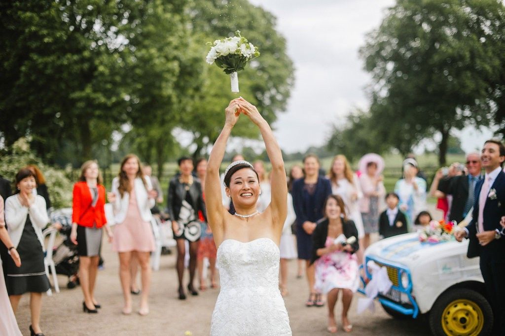 Joyful bride in strapless wedding gown tosses bouquet high into the air surrounded by eager wedding guests outdoors.