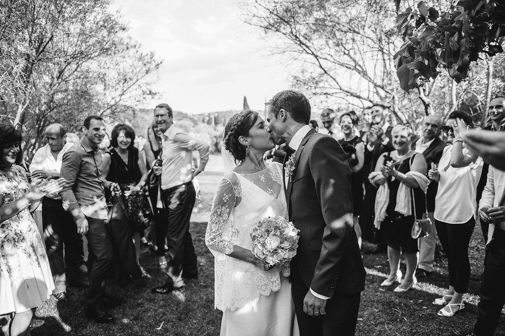 Black and white photo of newlyweds sharing a kiss while guests throw confetti at outdoor wedding.