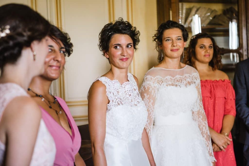 Two brides in white lace wedding dresses surrounded by smiling bridesmaids in pink dresses at reception.