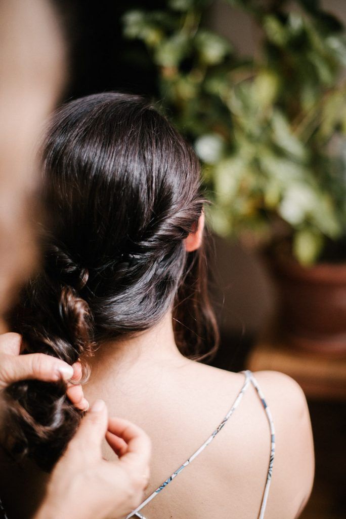 Hairstylist pinning elegant braided updo on dark-haired bride during wedding preparation.