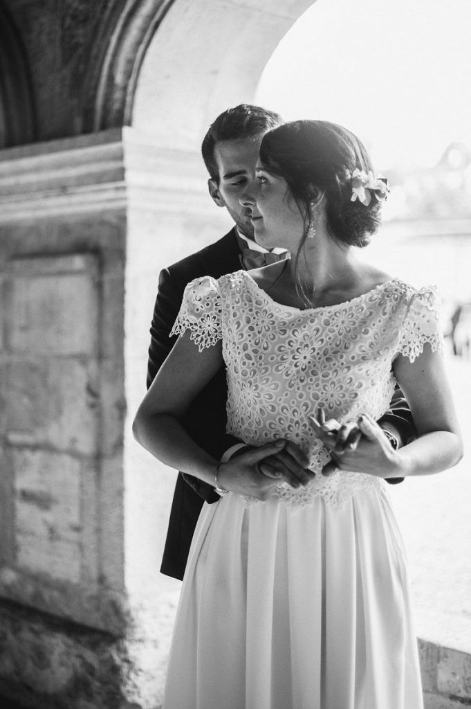 Groom tenderly kissing bride on the cheek under a stone arch in black and white.
