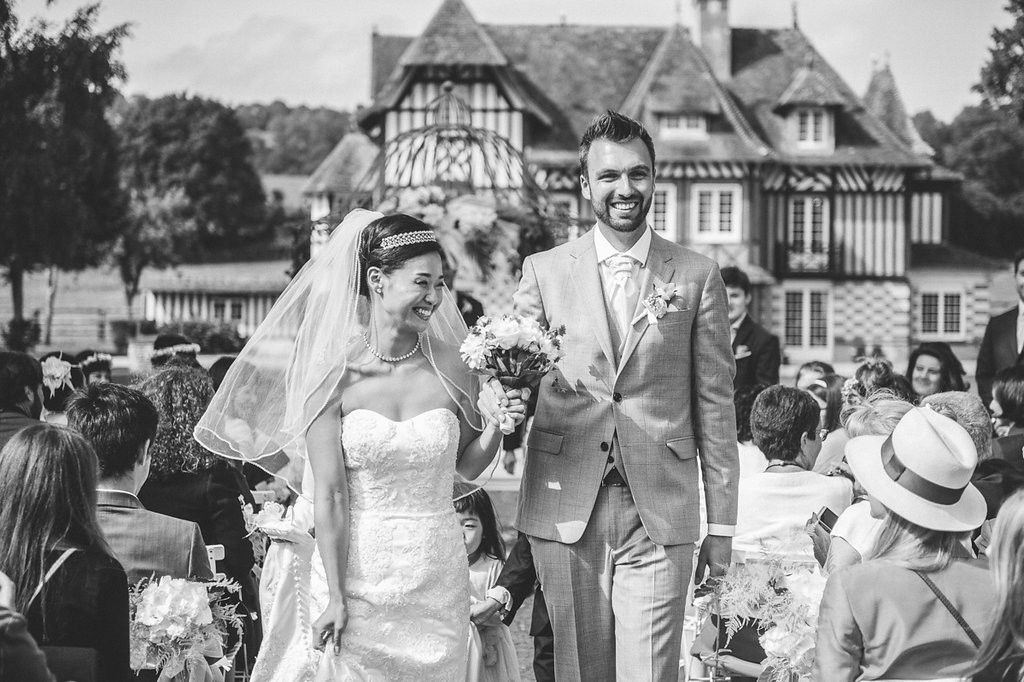 Newlywed couple walks joyfully down the aisle after wedding ceremony with a stunning French manor house backdrop.