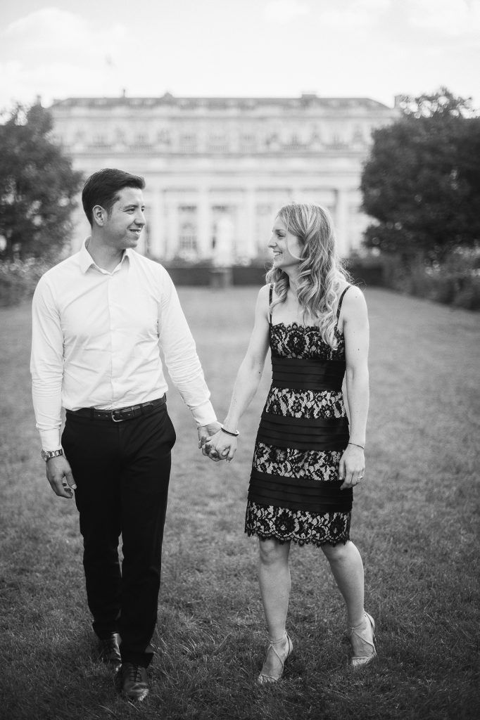 Couple holding hands and smiling at each other while walking on grass with grand building behind them.