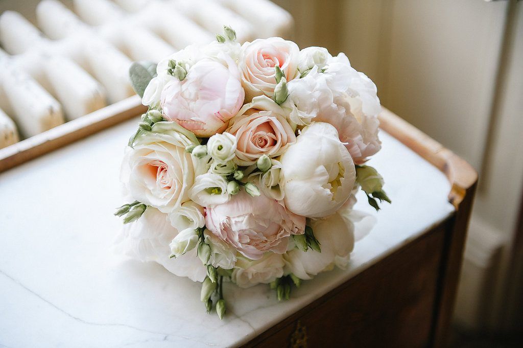Elegant bridal bouquet of blush peonies, cream roses, and white flowers on marble surface.