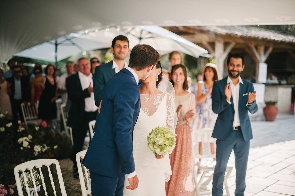 Newlyweds share first kiss as married couple while guests clap and celebrate at outdoor wedding.