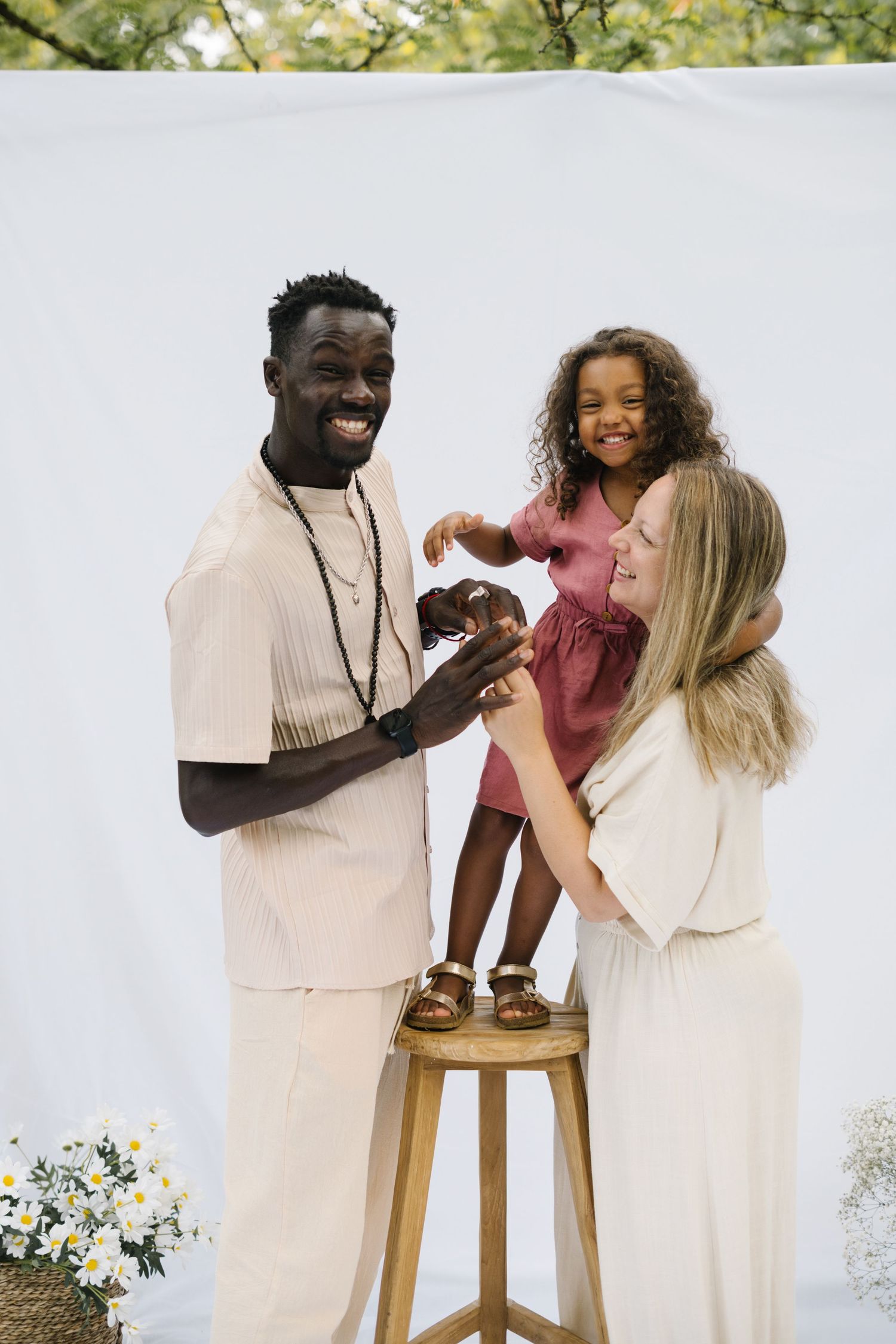Joyful interracial family with a young girl standing on a stool between her smiling parents outdoors.
