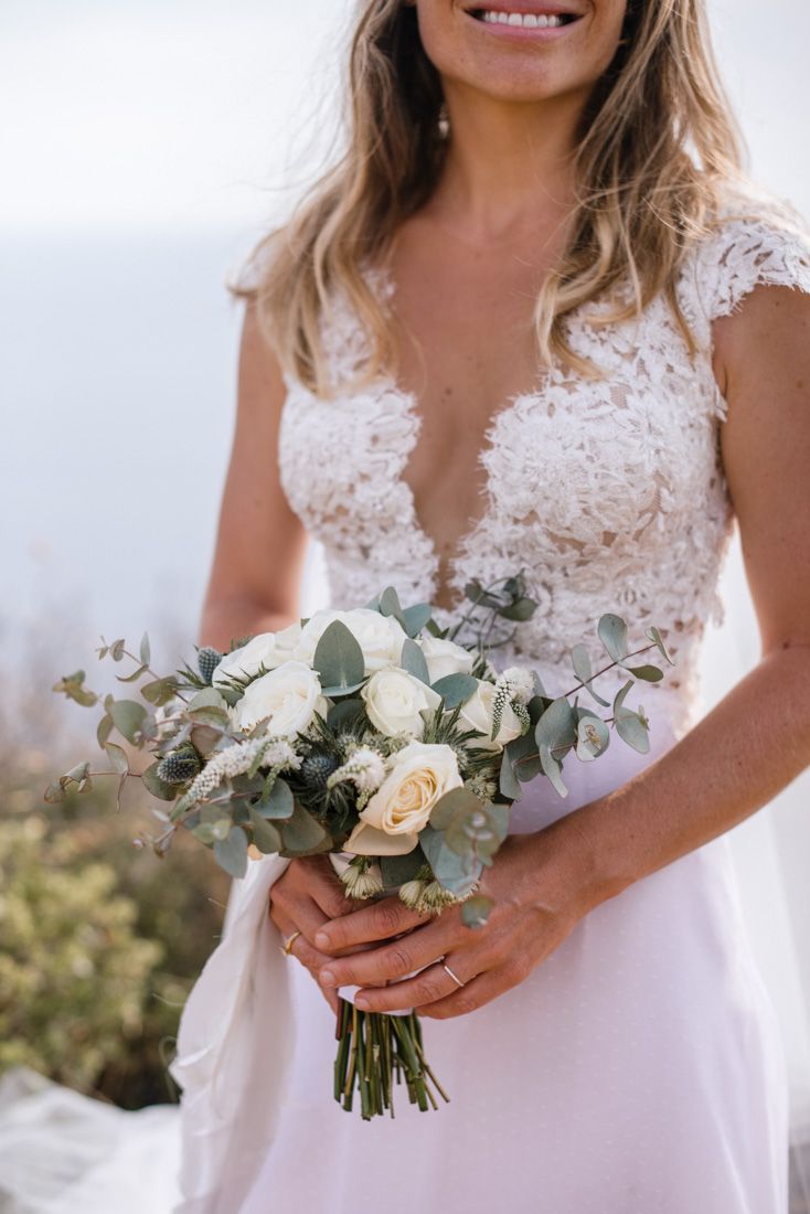 Bride in lace wedding dress holding elegant white rose and eucalyptus bouquet outdoors.