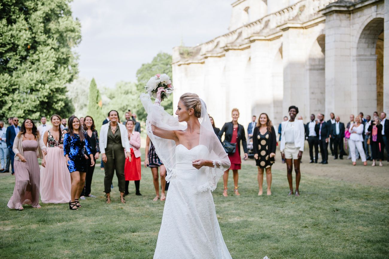 Bride tossing bouquet to wedding guests gathered outdoors near a classic stone building.