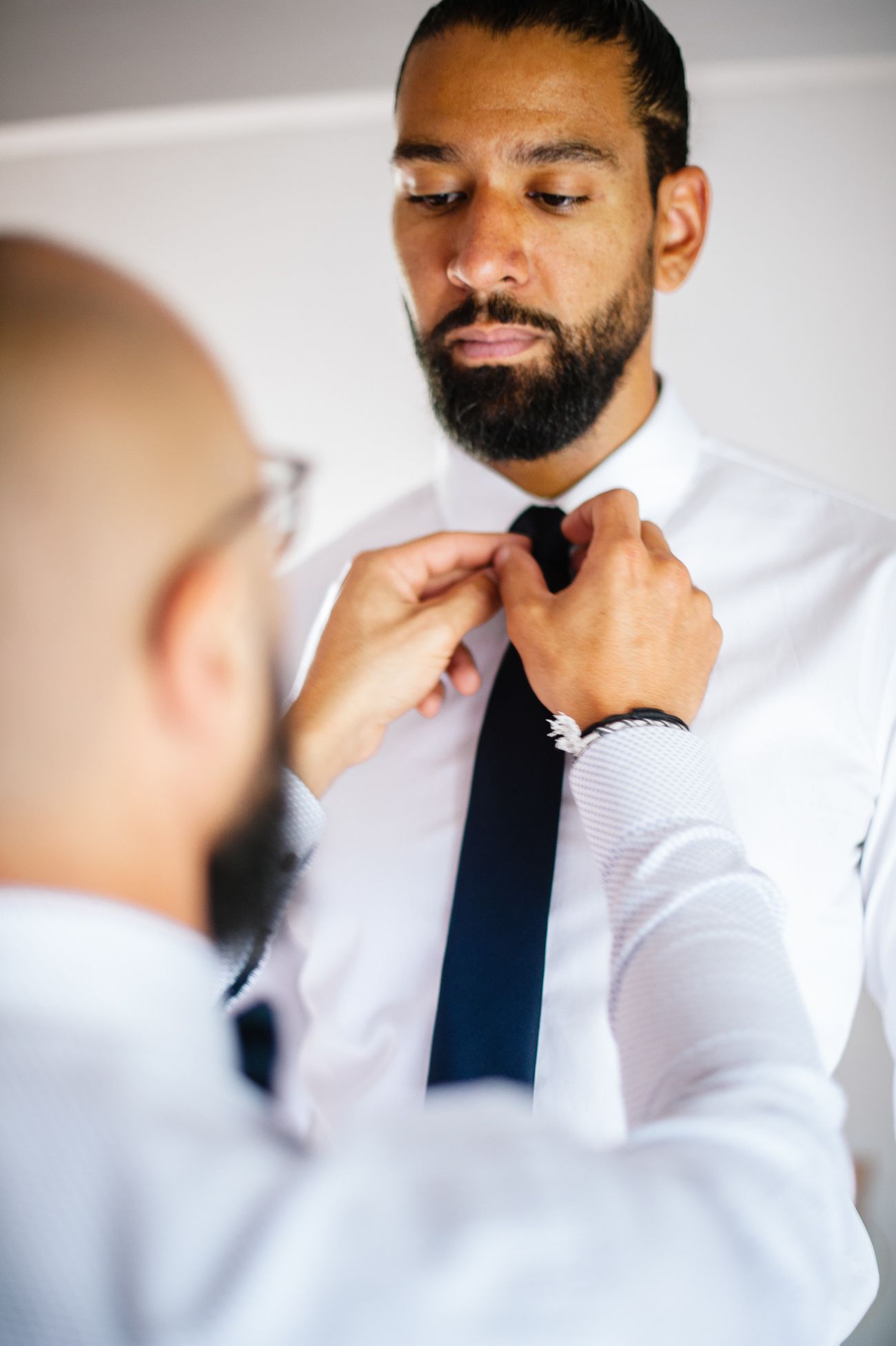 A man in a white shirt adjusts another man's navy tie during wedding preparations.